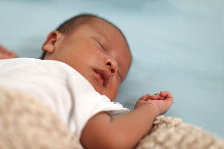 Sleeping newborn baby in a white onesie, resting on a soft beige blanket against a pale blue background.
