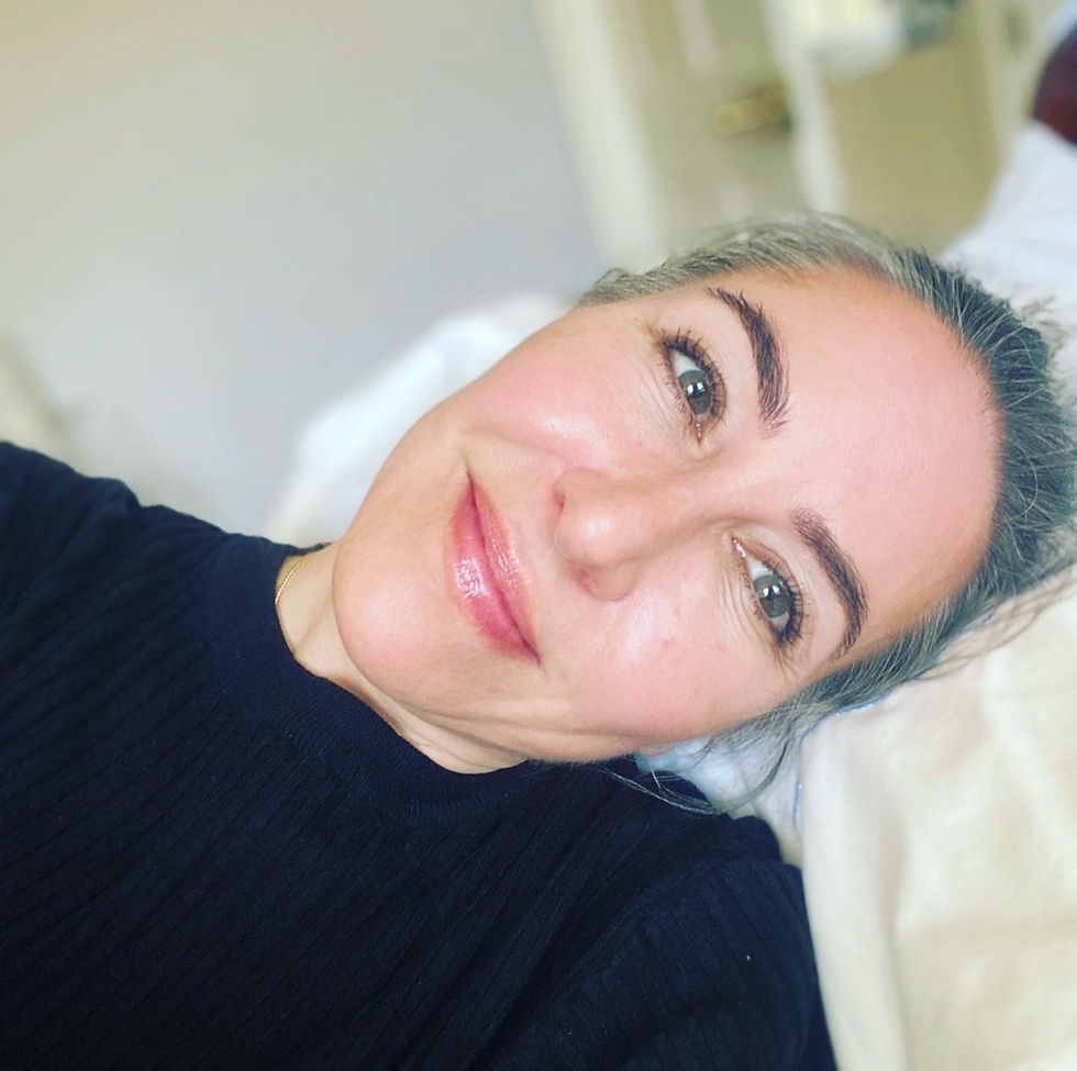 Smiling person lying on a bed, wearing a black top. Soft lighting creates a relaxed and content mood. Pale background. No text visible.