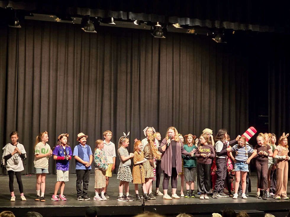 Children in animal costumes and casual clothes stand on a stage while a woman speaks into a microphone before black curtains.