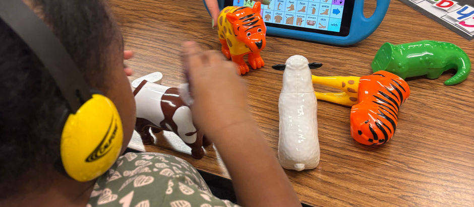 Child wearing ear defenders in a classroom points at an AAC tablet while colorful animal toys sit on a table, showing focused learning.