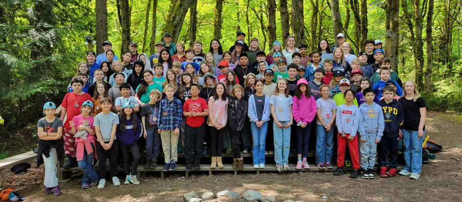 Large group of smiling kids and adults posing in a forest near a stone-ringed campfire.