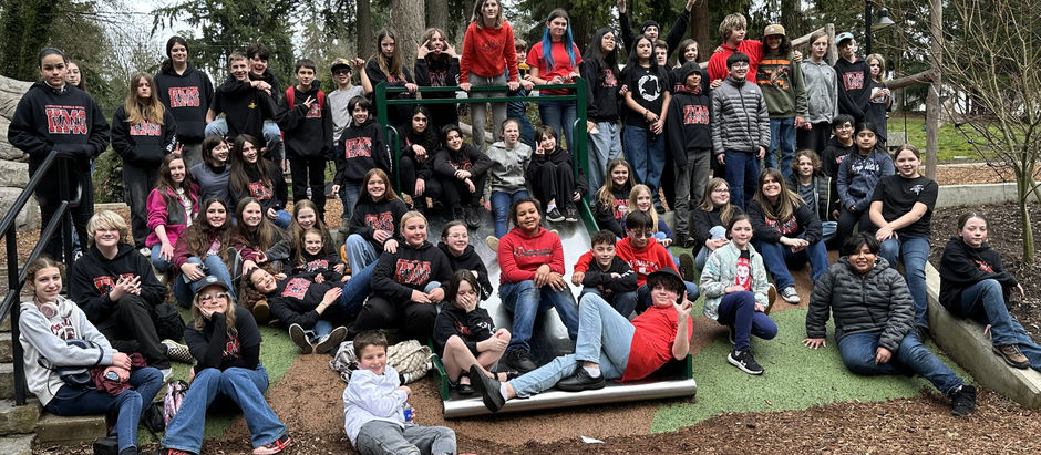 A large group of smiling kids in matching black hoodies gather around a slide in a park. Trees and cloudy sky are in the background.