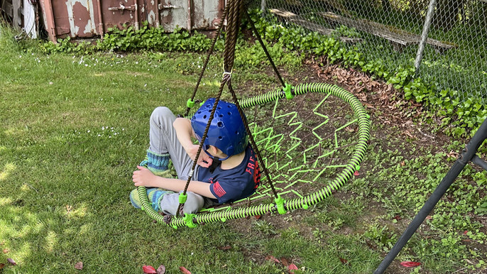 Child with a blue helmet sitting in a green rope swing in a garden. Wearing blue shirt, gray pants. Fence and shed in the background.