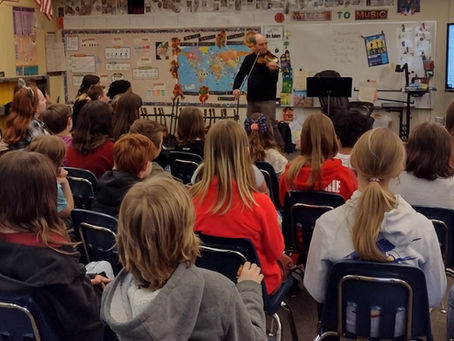 Children stand and smile in a colorful classroom as a man plays a violin. Musical notes and educational posters are on the walls.