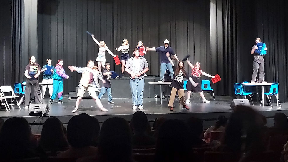 Performers on stage, dance energetically against a black curtain backdrop. Blue chairs and tables frame the scene. Actors perform Where do you belong? Choreography.