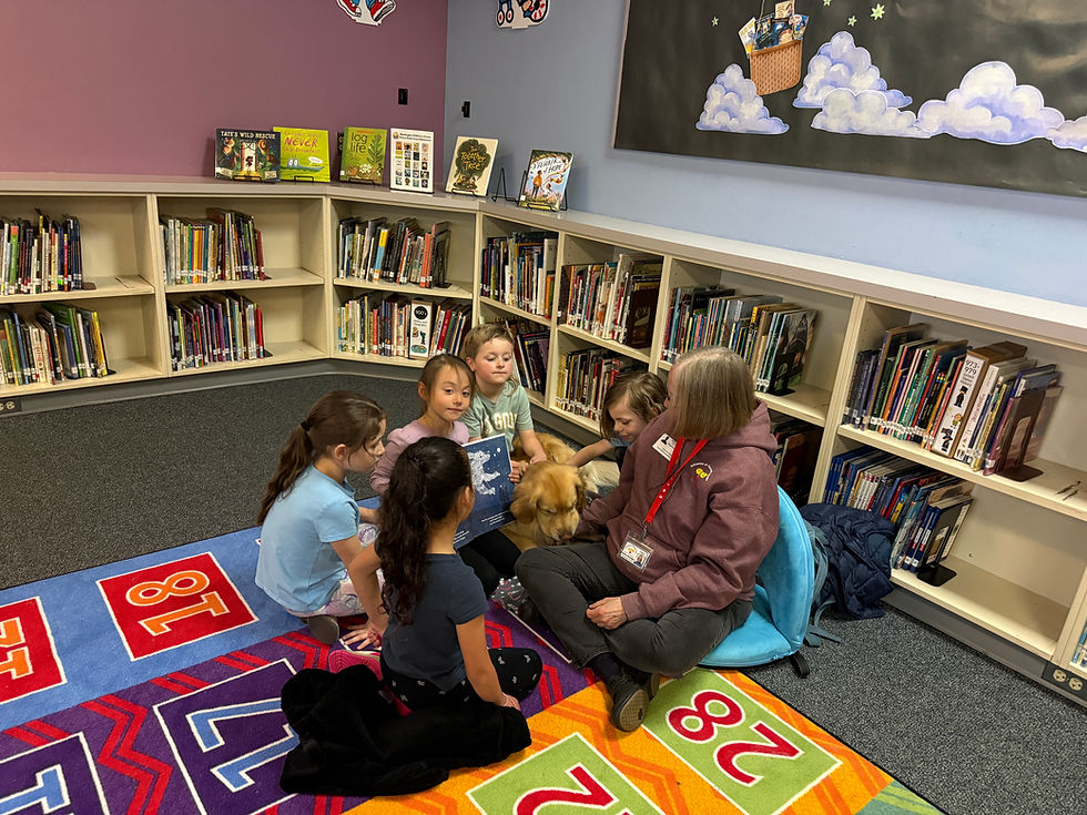 Children and a teacher sit on a colorful library rug petting a golden dog, surrounded by bookshelves and picture books.