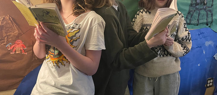 Three students standing and holding "The Hobbit" book infront of a wall decorated with colored paper showing scenes from the book.