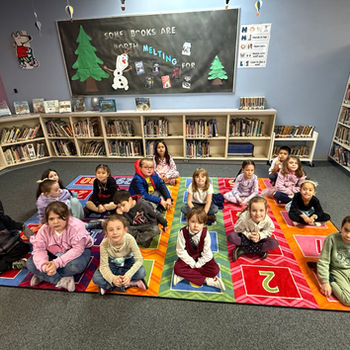 Children sit cross-legged on a colorful number rug in a school library, beneath a bulletin board reading Some books are worth melting for.