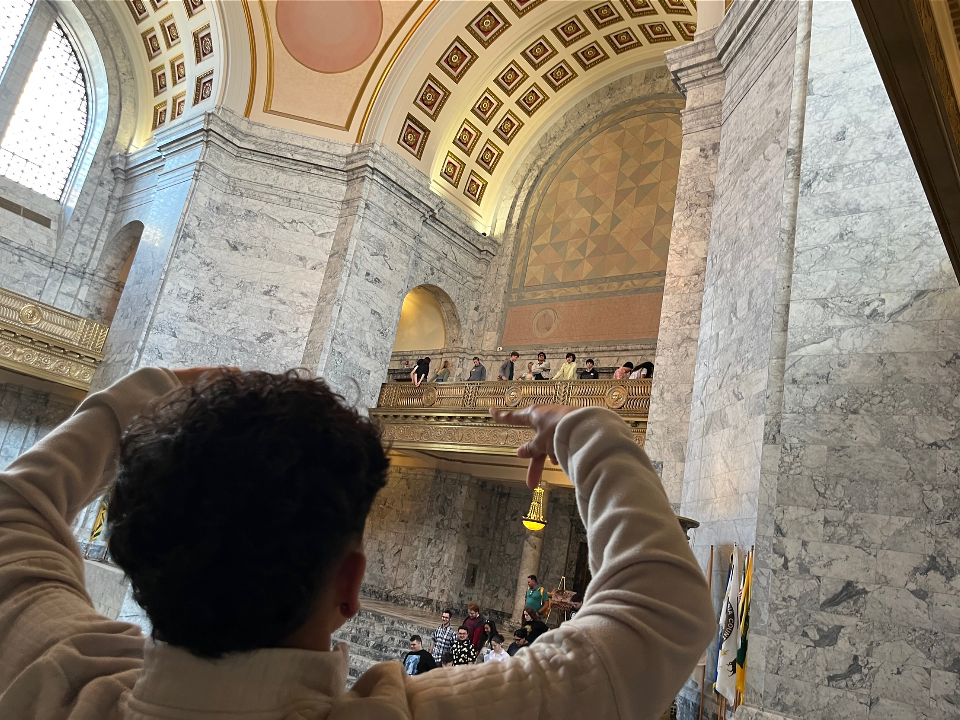 A highschooler getting the attention of his friends across a large arched room in the state capital building