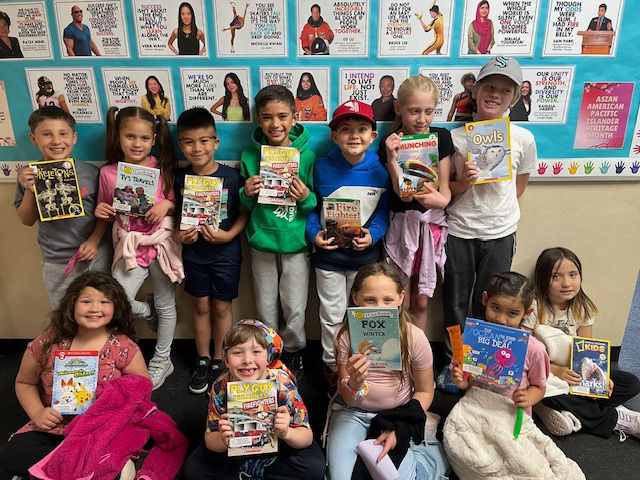 Children smiling and holding books in a classroom against a colorful bulletin board with images and text.