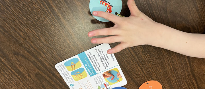 A child's hand spins a blue disc with a caterpillar on a wooden table. Instruction card visible, alongside blue and orange discs.