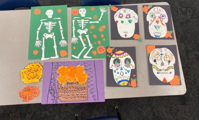 Student art collage of paper skeletons and decorated skull masks on a gray tabletop, in green, purple, black and orange.