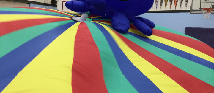 A large blue inflatable octopus with a smiling face rests on a colorful striped parachute in a gymnasium. Banners and hoops in the background.