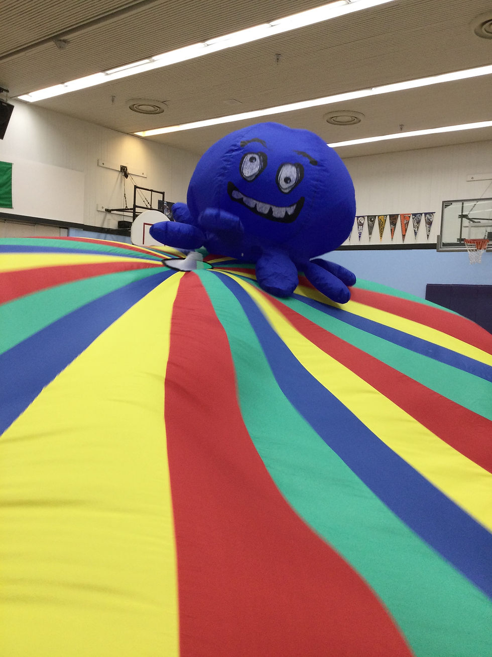 A large blue inflatable octopus with a smiling face rests on a colorful striped parachute in a gymnasium. Banners and hoops in the background.