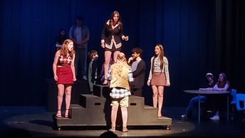 Theater scene with three actors in various poses on black steps. Multiple actors in background. Plastics scene in Mean Girls.
