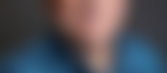 Dennis Peters smiling, wearing glasses and a blue patterned shirt, set against a dark gray background.