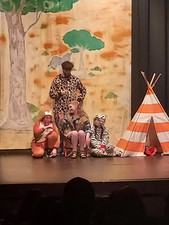 Mean Girls Africa scene. Three actors wear animal print onsies. The fourth actor sits in a chair surrounded by the actors. There is an orange striped tent beside them. An African painted scene in the background.