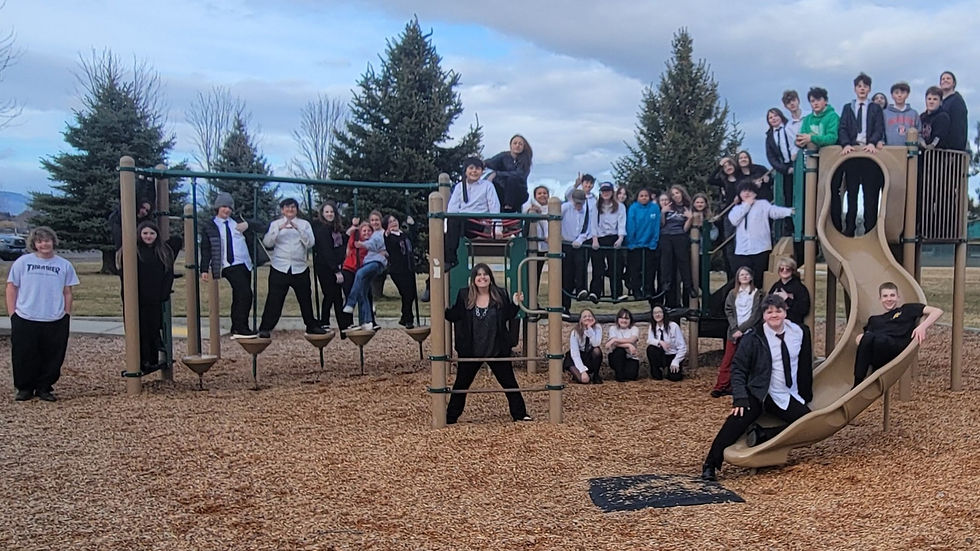 A large group of teens pose on a playground structure, some standing on bars and one sliding down. Overcast sky and trees in the background.
