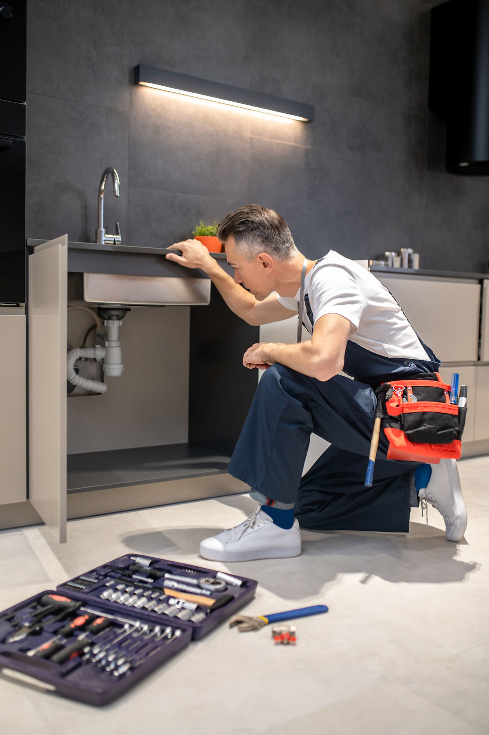 Plumber examining sink in kitchen, with toolbox and utility belt. Handyman Services