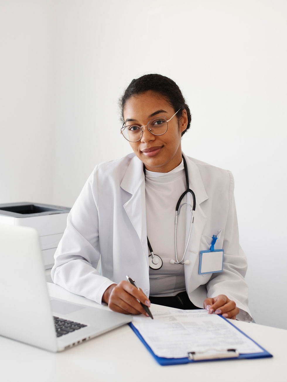 Smiling doctor writing on a clipboard, with stethoscope and laptop, Medical Care Centre.