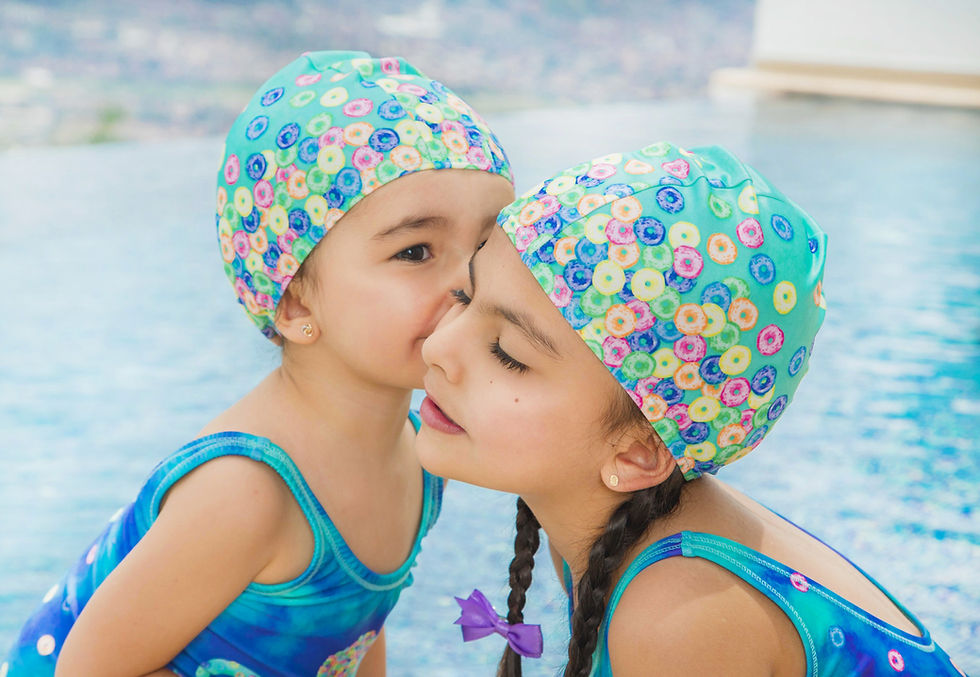 Thumbnail: dos niñas con vestido de baño entero en lycra de color azul y aguamarina ilustrado con pulpo de fruit loops de colors y con gorro en lycra de color aguamarina ilustrada con fruit loops de colores y al fondo piscina