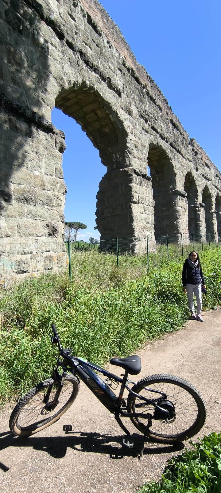 A bicycle in the foreground with Raquel standing in the distance near a majestic ancient Roman aqueduct at the Park of the Aqueducts in Rome