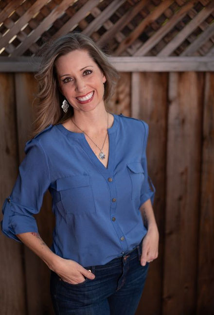 a blond woman smiling in a blue shirt and jeans standing in front of a fence