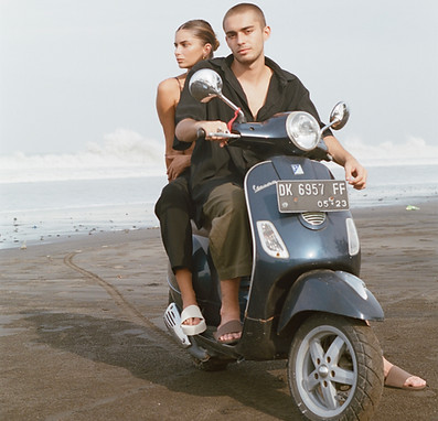 Lifestyle fashion image of a man and woman sitting on a blue-grey motorcycle, wearing dark linen outfits and Indosole sustainable sandals in white and brown.