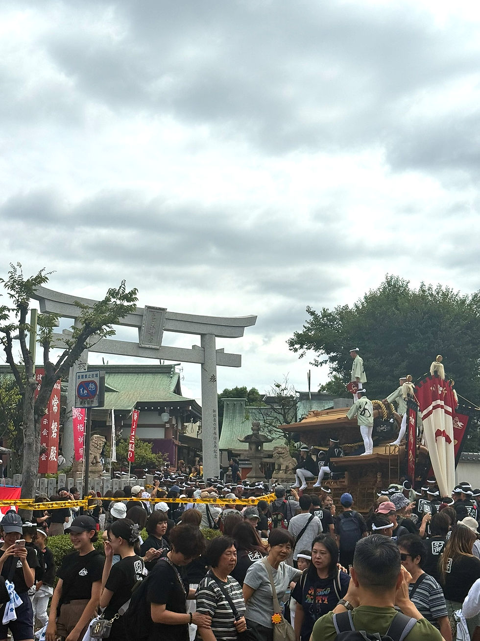 Crowd gathers at a Japanese shrine festival under cloudy skies, watching men in white and black atop a wooden float by a torii gate.