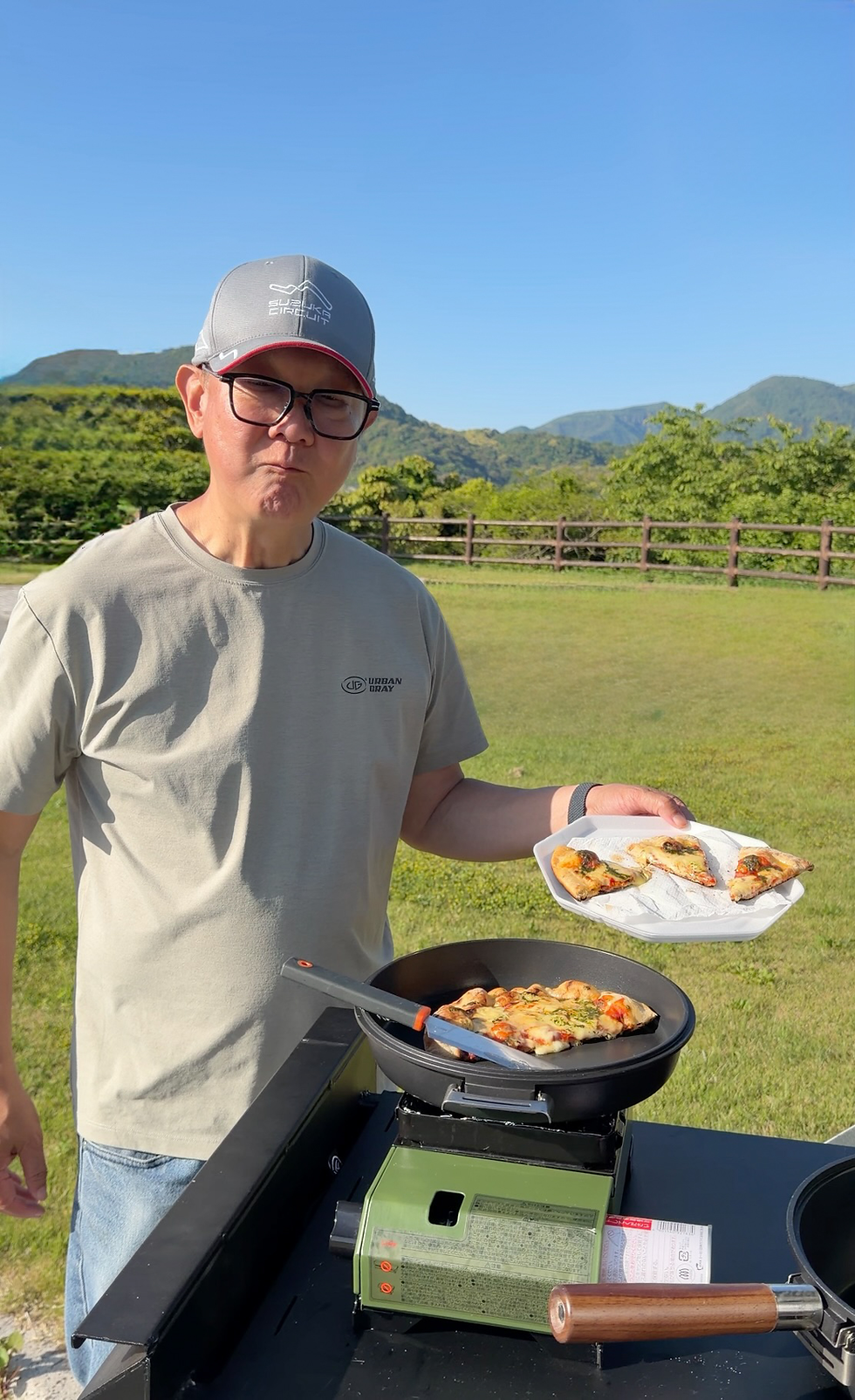Man in cap and glasses serves pizza slices from a pan outdoors, with mountains, green field, and blue sky behind him.