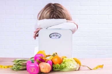 A woman holds a scale behind dumbbells and a pile of nutritious food.