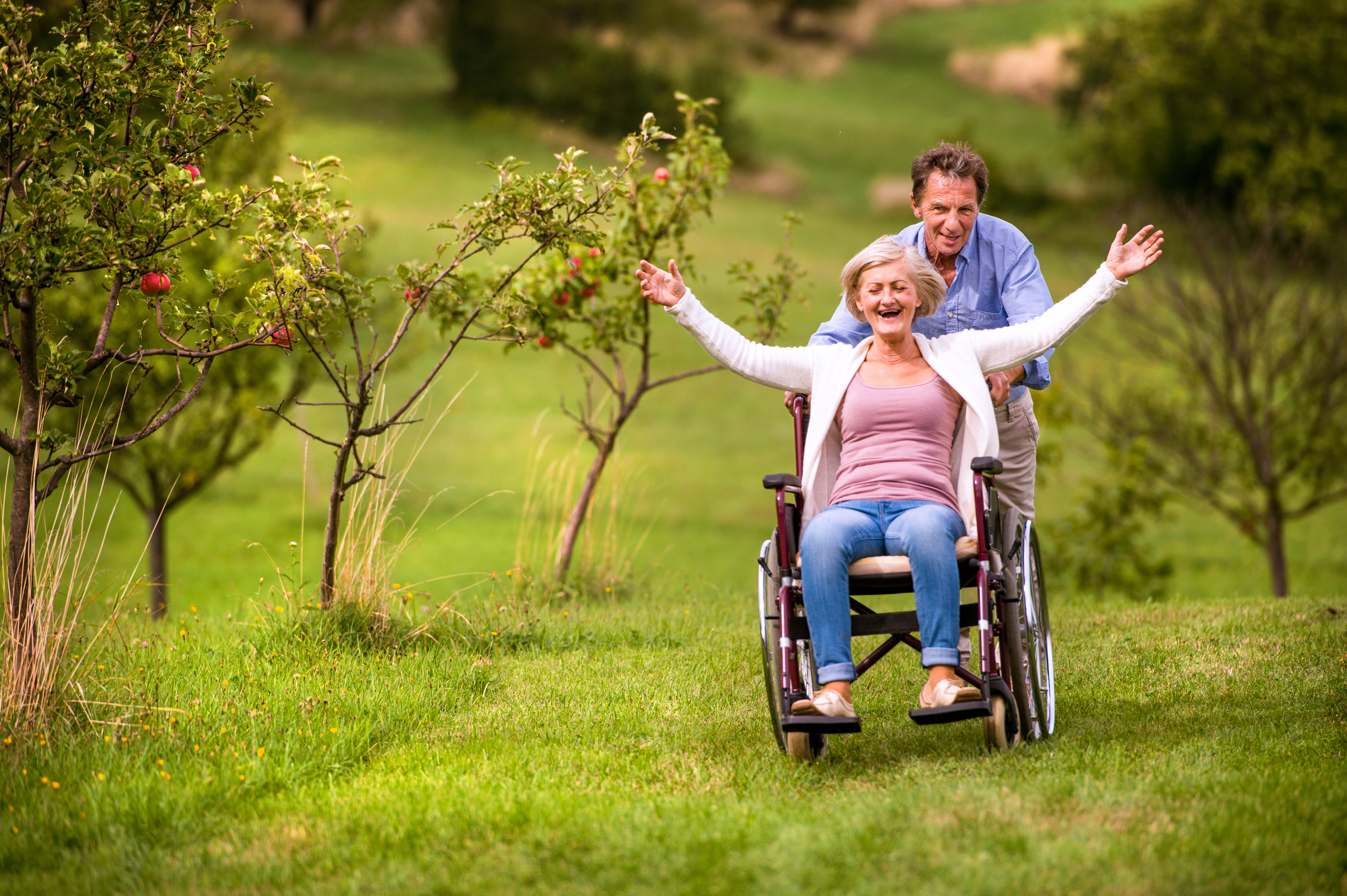 Senior man pushing a smiling woman in a wheelchair outdoors in a green park