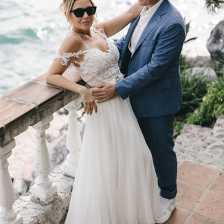 An elegant couple in a romantic embrace by Lake Atitlán; the bride is wearing a white dress and sunglasses, and the groom is wearing a blue suit. 