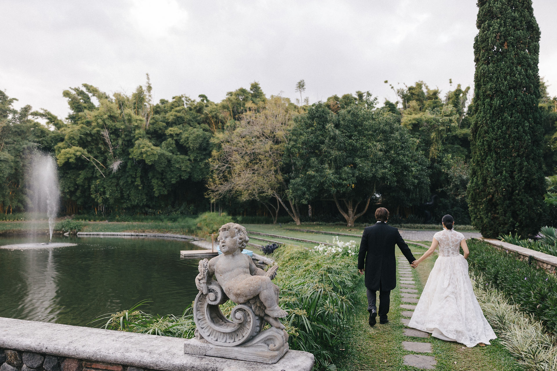 A newlywed couple walking hand in hand along a path in a lush garden featuring a pond, a fountain and a cherub statue at Villa Bokeh, Antigua Guatemala.