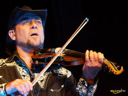 Cowboy-hatted violinist, Geoffrey Castle, in embroidered black shirt plays on a dark stage, focused under blue-orange light.