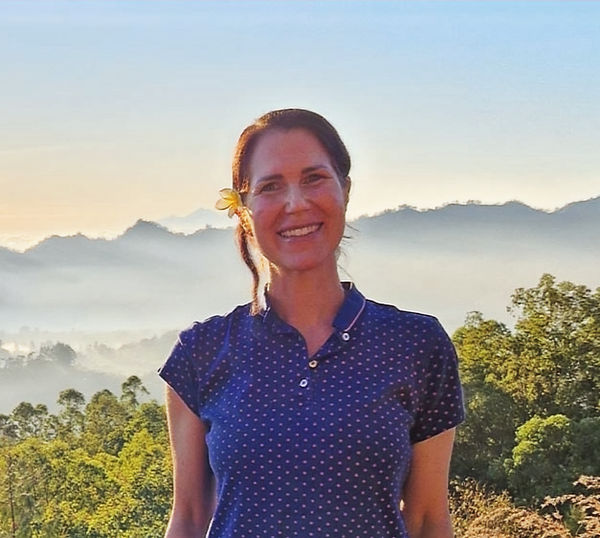 Ein Foto von Sylvia. Sie lächelt freundlich. Im Hintergrund ist eine tropische Landschaft zu sehen.