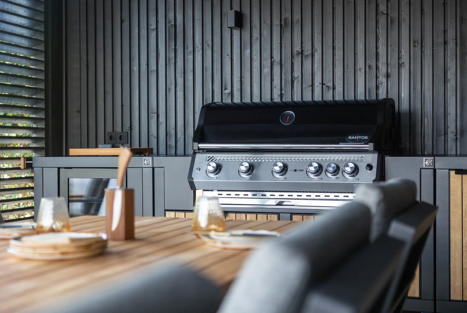 Cuisine extérieure Rubix Duo Roostr intégrant un barbecue gaz Santos, modules anthracite et façades en lames de bois, installée sous abri près de la table à manger.
