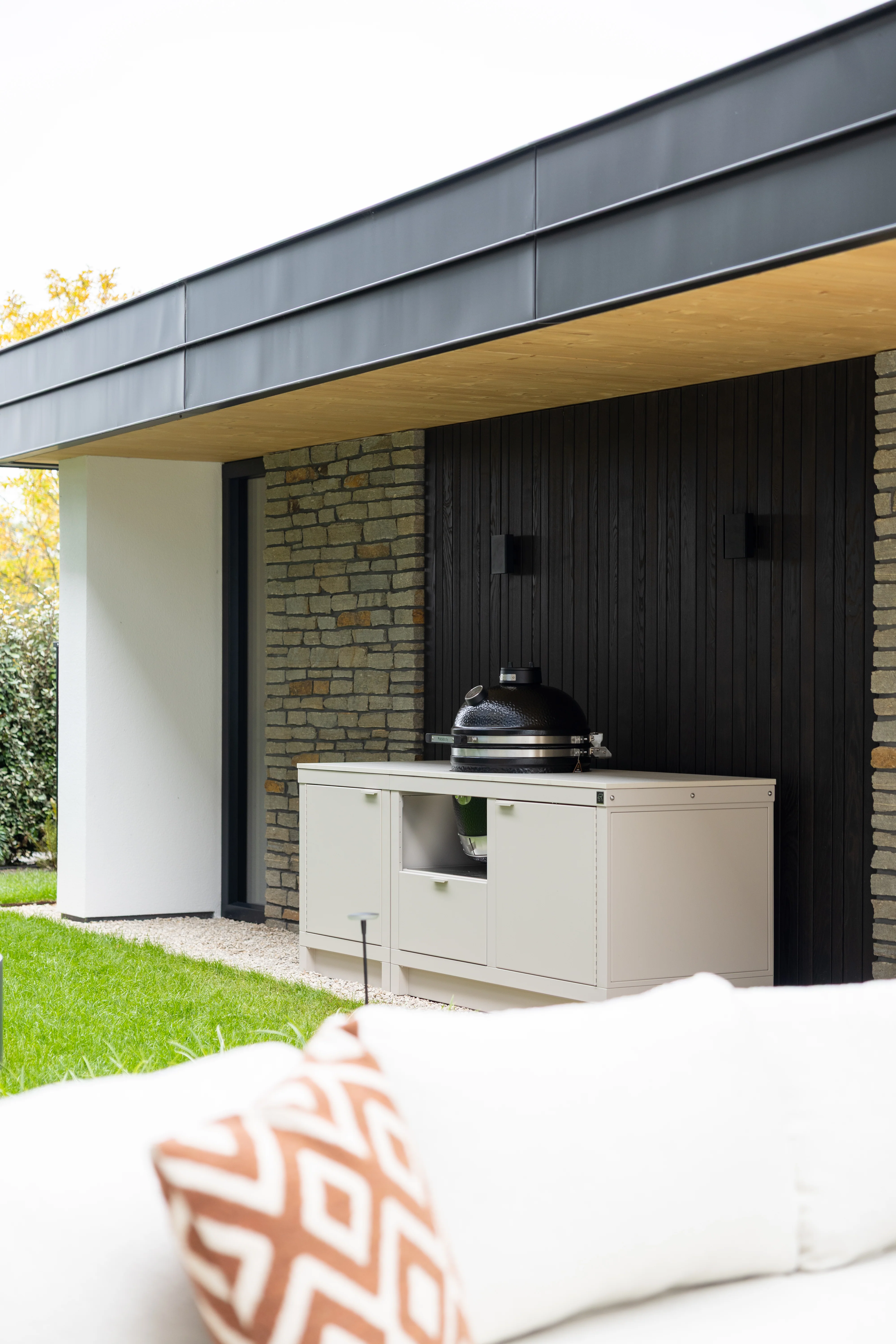 Cuisine extérieure Rubix couleur sable avec Kamado intégré, installée sous auvent contre bardage noir et mur en pierre – style contemporain WAHO x Roostr.