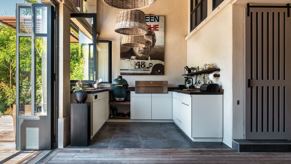 Cuisine extérieure sur-mesure avec meubles encastrés, plan de travail en bois, barbecue haut de gamme et kamado, intégrée dans un espace couvert au design chaleureux.