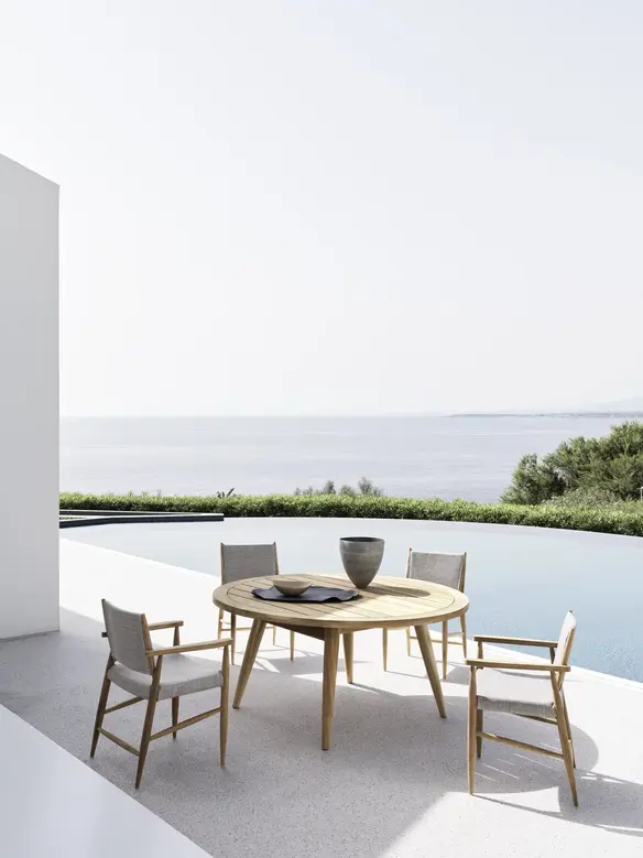 Table ronde Eidos en teck installée sur une terrasse surplombant la mer, entourée de 4 fauteuils.