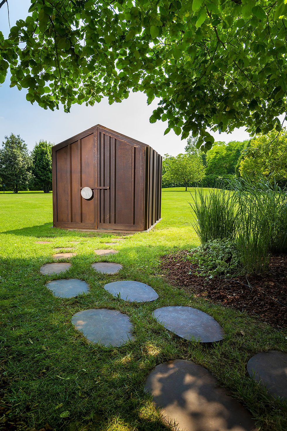 Cabane « La pièce qui n’existe pas » d’Il Giardino di Corten en acier corten patiné, façade brute avec serrure artisanale, posée sur pelouse ; pas japonais menant à la micro-architecture.