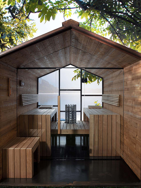 Intérieur version sauna – Banquettes en bois et poêle central dans La pièce qui n’existe pas, grande baie vitrée ouvrant sur le paysage.