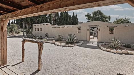 A view from inside the shooting range area at the Mas de Campe property, showing a rustic wooden structure reminiscent of an old Western town. The area is surrounded by desert-style landscaping with various cacti and succulents