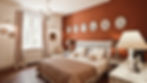 beautifully decorated bedroom in a villa located in the Alpilles mountain range, available for rent. The room features a cozy double bed with a white wicker headboard, adorned with soft pillows and a patterned quilt. The warm terracotta wall adds a touch of rustic charm, complemented by whimsical framed illustrations above the bed. Elegant bedside tables with ornate lamps enhance the room's sophisticated ambiance. A chandelier adds a classic, luxurious touch, while natural light streams through a large window, enhancing the bright and airy atmosphere. An inviting armchair in the corner provides a comfortable spot to relax. This bedroom beautifully blends authentic Provençal decor with a high level of comfort and luxury, making it an ideal retreat for guests seeking a serene and stylish stay in the Alpilles