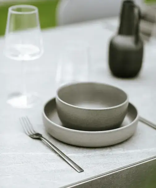 Détail d’une table dressée sur le plan de travail de la cuisine extérieure Fogher AGHER, avec vaisselle design et verre à vin sur fond de verdure.