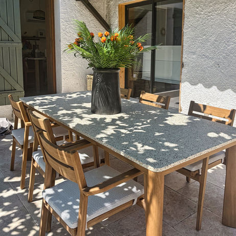Espace repas extérieur avec table rectangulaire Talenti Oliver, chaises assorties en teck et coussins clairs, décoré d’un vase noir Gommaire GINGER garni de fleurs oranges, sous une pergola ombragée.