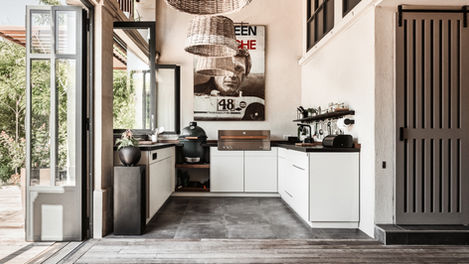 fully equipped outdoor kitchen with large windows opening to a deck and garden. The space includes a ceramic barbecue, gas grill, dishwasher, induction cooktop, and a refrigerator drawer, all designed for a luxurious outdoor cooking experience