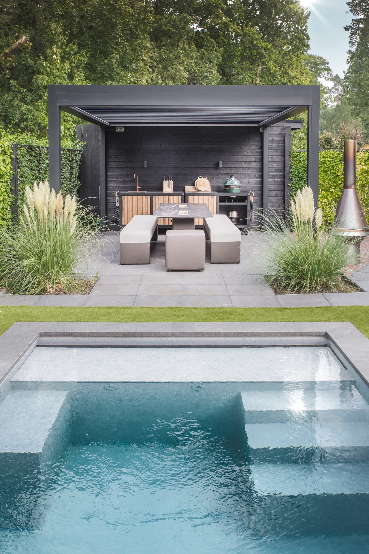 Espace repas avec pergola et cuisine Rubix : façades bois ajourées, évier cuivre et Kamado Big Green Egg intégré face à la piscine.