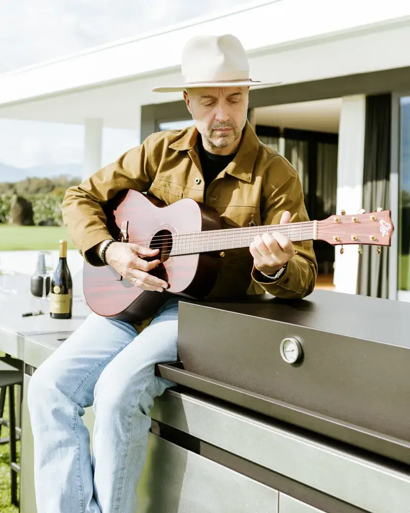 Joe Bastianich jouant de la guitare sur la cuisine extérieure AGHER Fogher, dans une ambiance détendue en plein air, avec une bouteille de vin à ses côtés.