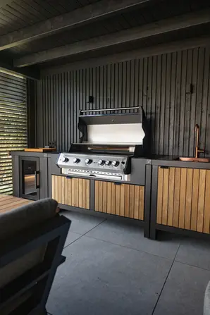 Cuisine d’extérieur Roostr avec grand barbecue à gaz intégré, réfrigérateur et évier cuivré, sur fond de bardage en bois noir